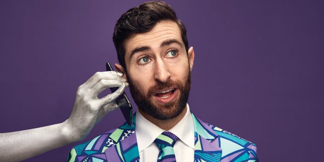 A silver hand holds a phone up to the ear of a bearded man in a colorful suit, against a purple background.