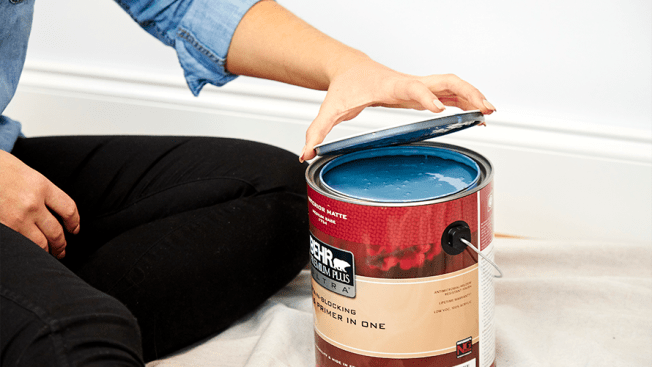 A hand lifting the top off a can of blue Behr Paint