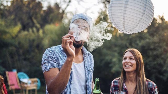 there is a couple in the image; the man holds a beer in one hand; in the other hand the man holds a cannabis cigarette and exhales a large cloud of smoke