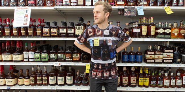 A man wearing a bourbon camouflage shirt stands in front of a shelf of bourbon.