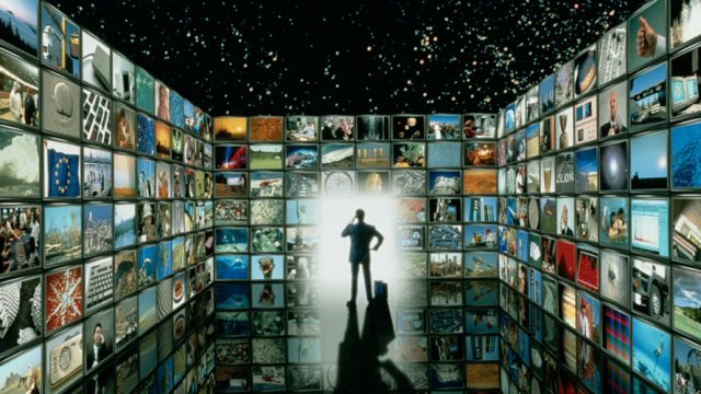 A man is standing; around him are screens on each side of him; on the massive screens there are pictures from throughout history like the moon landing