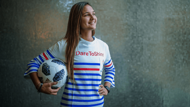 Female Soccer star poses with a soccer ball in a red, white, and blue striped shirt that says 