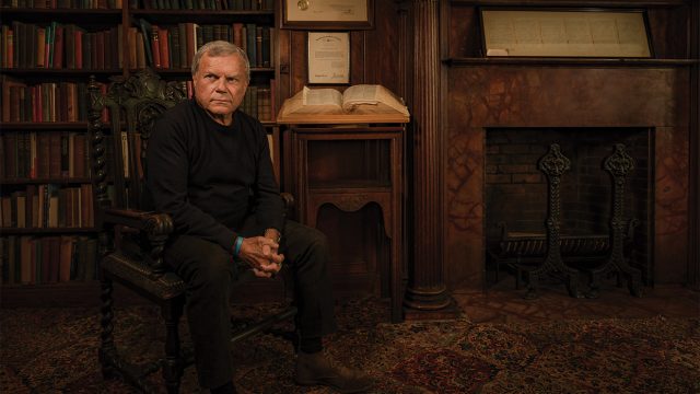 Martin Sorrell sitting in a chair in a library in front of a bookcase, a large open book and a fireplace.
