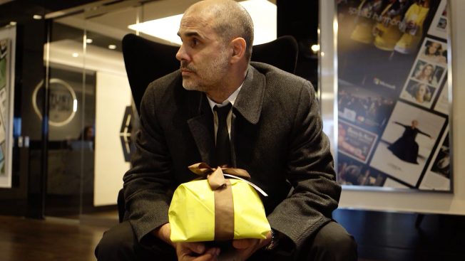 Gerry Graf wears a shirt, tie and coat and sits in a chair, holding a gift wrapped in yellow with a brown bow.