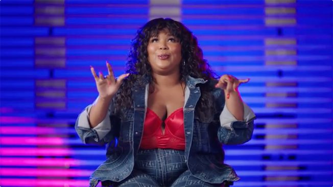 Lizzo smiling in a denim jacket and red corset on a blue background