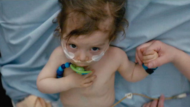 A toddler with a pacifier lies in a hospital bed and holds hands with an adult