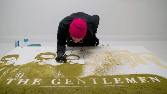 An artist bends over a poster made of cannabis with the words The Gentleman