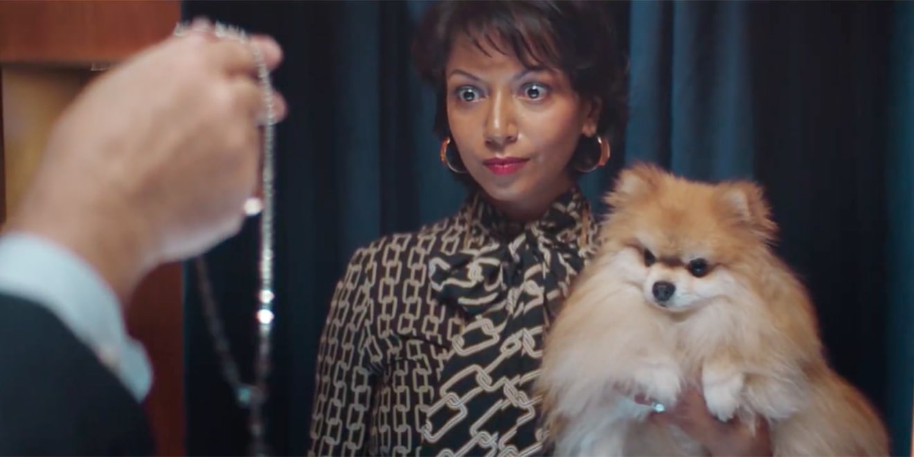 A woman with wide eyes holding a dog and looking at a man's hand holding a piece of jewelry
