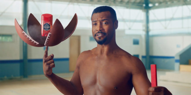 Isaiah Mustafa using his index finger to hold up a football that's open with Old Spice in the middle