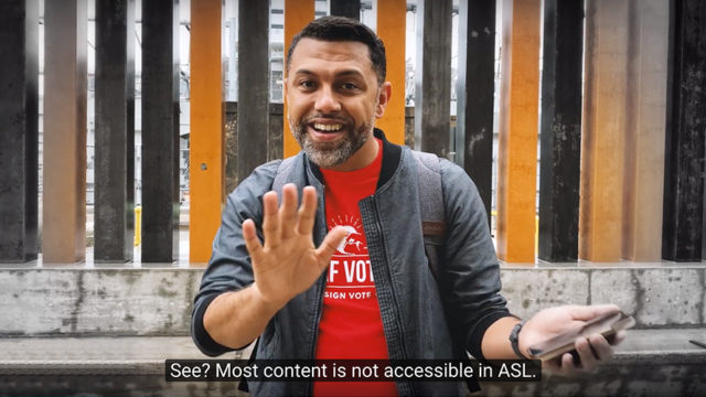 A man with a beard and dark hair, wearing a red t-shirt and light jacket, stands on the street speaking ASL. The caption reads: 