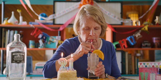 Arlene Manko drinking an alcoholic beverage next to a birthday cake with 2 and 1 candles and a bottle of Aviation Gin