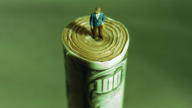 a tiny man standing on a giant roll of $100 bills