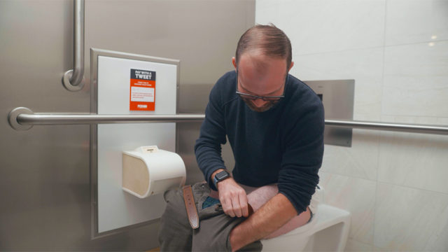 A man on a toilet fumbles for change in his pocket to pay a coin-operated toilet paper dispenser