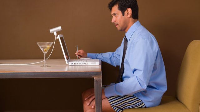 a man sitting at a desk in his underpants with a button-down shirt on and a martini next to his laptop