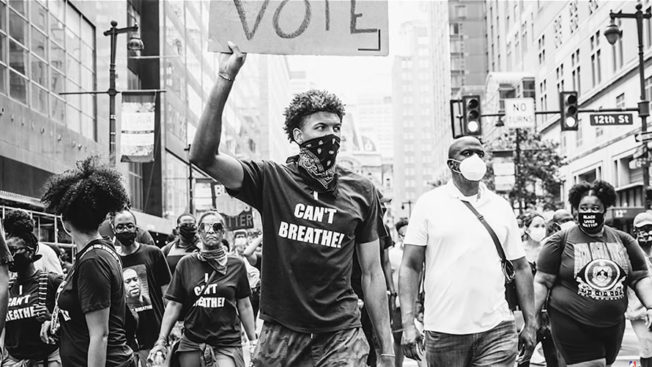 black and white photo of people protesting wearing masks with someone holding a sign that says vote