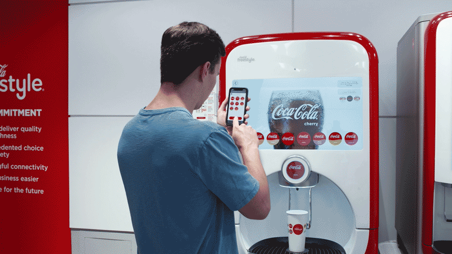 a man using his phone to select a coke from a coke vending machine