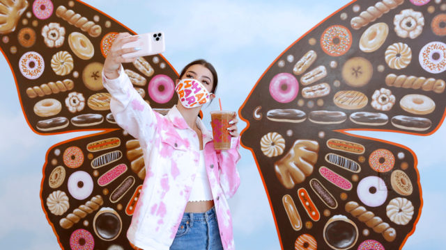 a woman standing in front of a mural of butterfly wings in a pink mask taking a selfie with a dunkin' cup