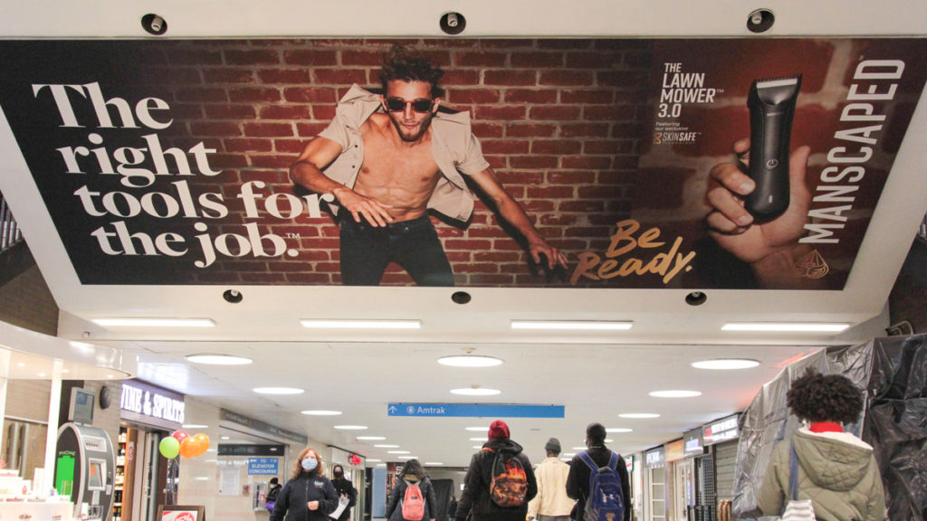 a man running with a caption: The right tools for the job on the left, and Be ready and the Manscaped logo on the right as people walk underneath the massive ceiling ad.