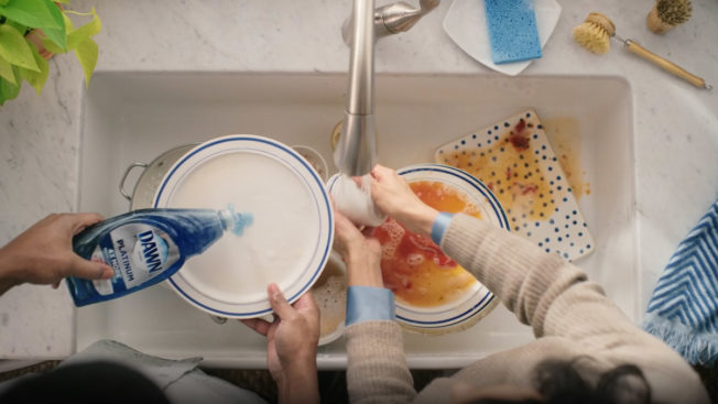 an overhead shot of hands cleaning dirty dishes in a sink with blue dawn dish soap