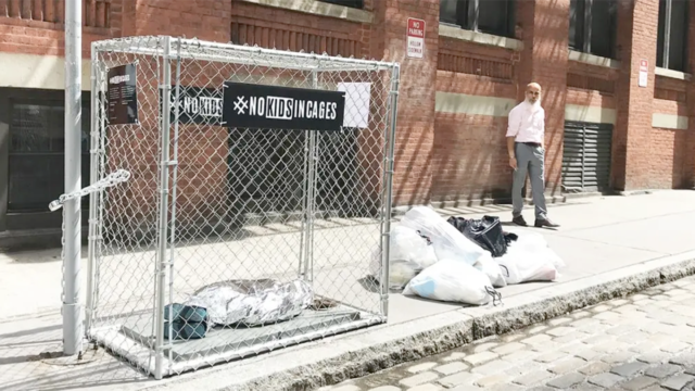 a man standing next to a cage that says #NoKidsInCages with what looks like a kid sleeping in the cage