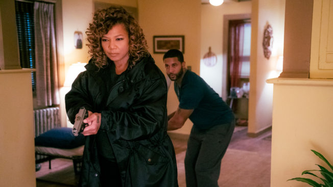 queen latifah holding a gun and walking in a room with a man behind her