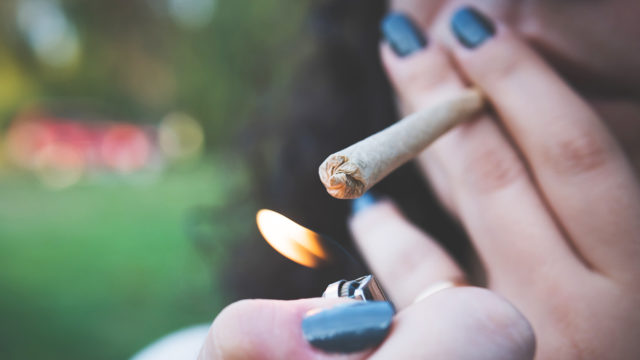 A woman with dark fingernail polish lights a cannabis cigarette in front of a blurred forested background