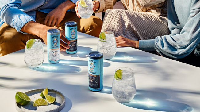 bombay sapphire gin and tonic cans on a table amid plates with hands in the background