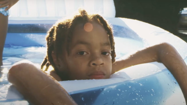 A young Black boy sits in an inflatable pool with his arms draped over the side as he looks toward the camera