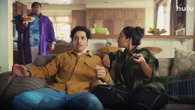 two people on a couch eating and sitting talking to someone behind them eating a pizza