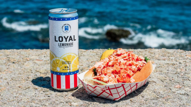 a loyal 9 drink next to a basket of a lobster roll in front of wave crashing