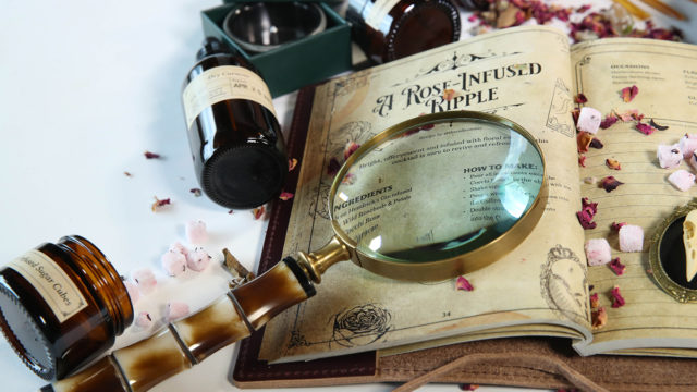 An antique style magnifying glass lies across a weathered page of a book seemingly filled with recipes and surrounded by bottles of ingredients