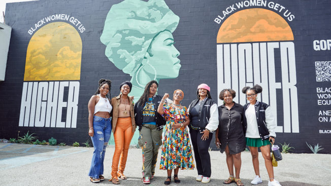 Women stand in front of a large mural that says 'Black Women Get Us Higher'