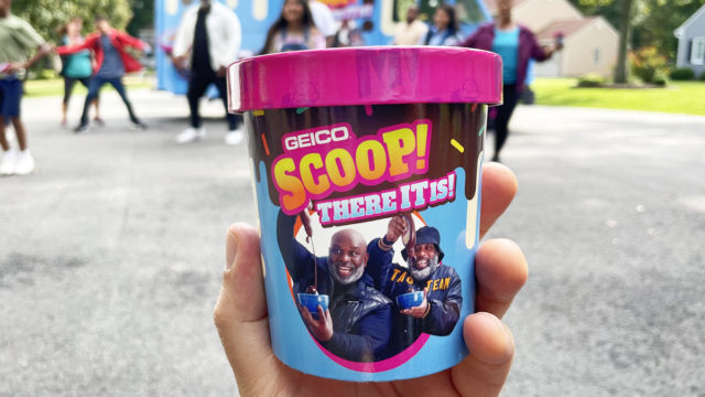 A hand holds a pint of Geico's Scoop, There It Is ice cream while people stand in the background