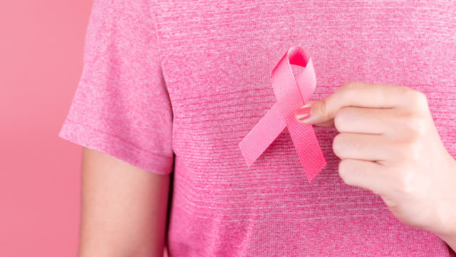 Photo of person in pink against a pink background holding a Breast Cancer ribbon close to their chest.