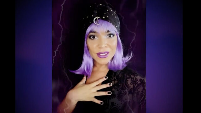 A woman with purple hair is dressed as a fortune teller in front of a dark background