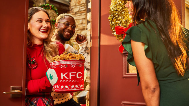 A woman at the doorway of a holiday party holds a KFC bucket encased in a knitted holder