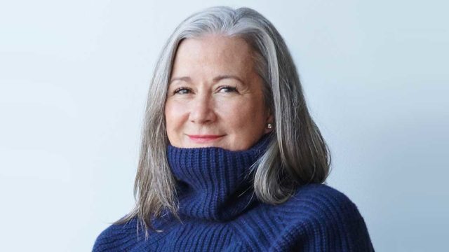Colleen DeCourcy is shown with shoulder-length silver hair and a blue sweater with high turtleneck collar