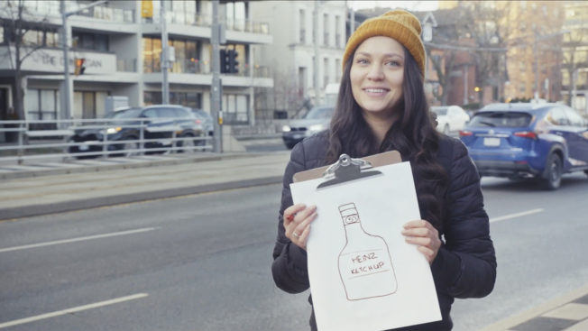 A woman in a yellow winter hat and dark jacket holds a picture of her amateurish drawing of a Heinz bottle