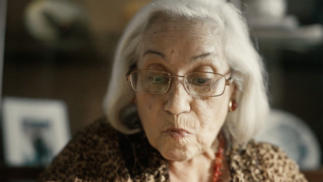 An elderly woman with shoulder-length gray hair looks down at her food while wearing glasses