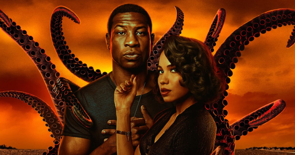 A man and a woman look into the camera while standing in front of tentacles and an ominous sunset