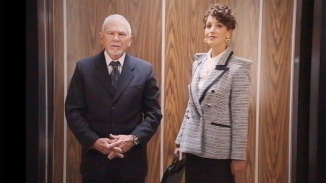 Two business people wearing 1980s-era work clothing stand in a wood-paneled elevator