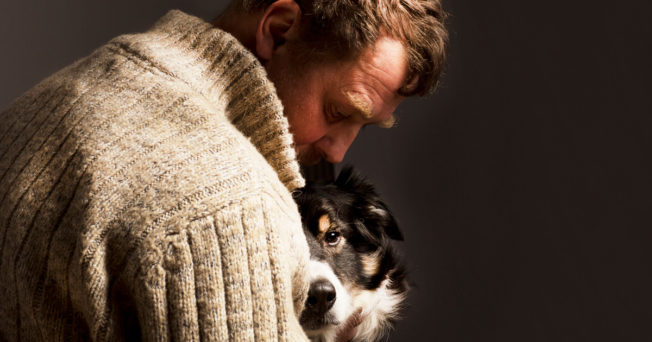 Man in white sweater hugging a dog.