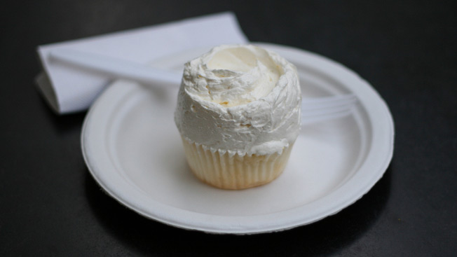 A white vanilla cupcake on a white plate with a white plastic fork and white napkin.